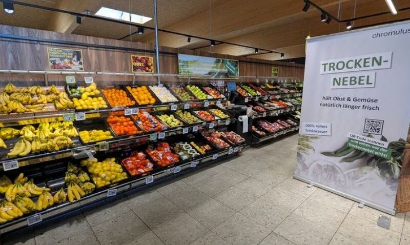 Ein Supermarktgang mit einer bunten Auswahl an Obst und Gemüse, darunter Bananen, Paprika und Äpfel, ordentlich in Regalen angeordnet. Rechts steht ein Werbebanner mit der Aufschrift 'TROCKEN-NEBEL' und Informationen zur Frischeverlängerung von Obst 