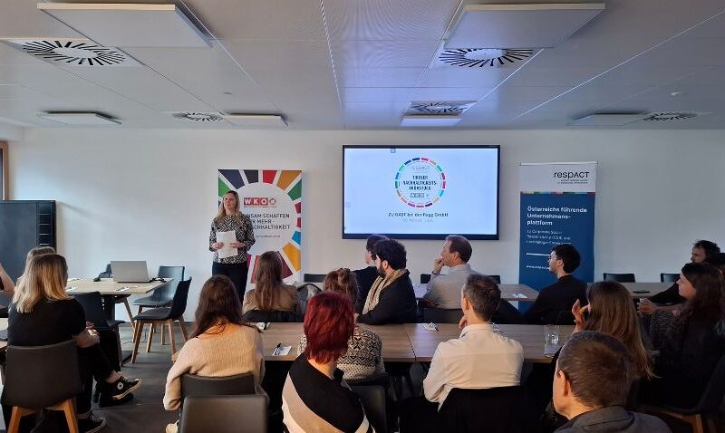 Eine Person hält einen Vortrag in einem Konferenzraum vor einer Gruppe von Menschen, die aufmerksam zuhören; im Hintergrund sind ein Bildschirm mit einer Präsentation und bunte Banner zu sehen.