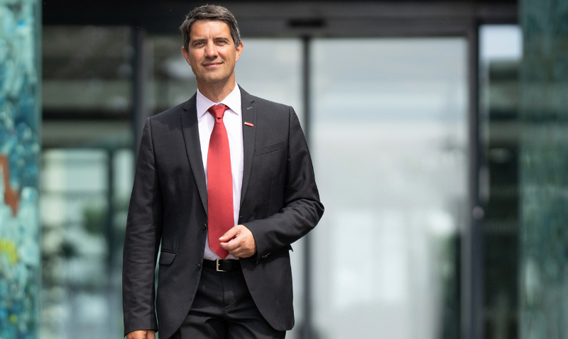 Mann im schwarzen Anzug mit roter Krawatte vor einem modernen Bürogebäude mit Glastüren